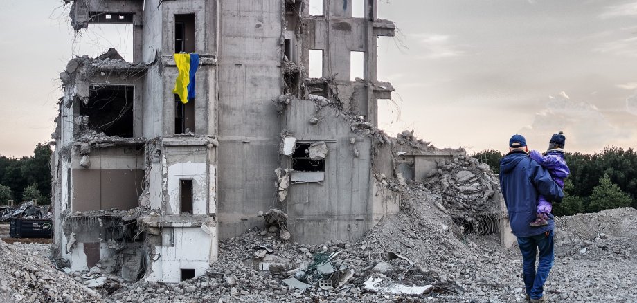 Mann mit einem Kind auf dem Arm vor einem zerstörten Gebäude